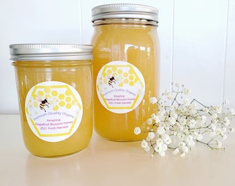 Puede incluir: Dos tarros de vidrio de miel con tapas plateadas. Los tarros están etiquetados con "Premium Quality Organic Amazing Grapefruit Blossom Honey 2021 Fresh Harvest". Un ramo de aliento de bebé blanco está en primer plano.