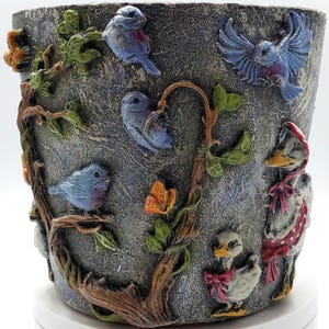 May include: A decorative planter with a textured gray finish, adorned with raised relief sculptures of bluebirds, butterflies, and ducks. The birds perch on brown branches with green leaves. The ducks wear red bows and bonnets.