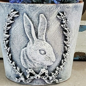 May include: A white ceramic planter with a raised design of a rabbit and a wreath. The planter has a distressed white finish.