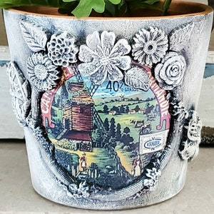 May include: A white ceramic flower pot with a distressed finish. The pot is decorated with a vintage label featuring a windmill and the text "40% Gras de Campagne". The pot is also decorated with 3D floral embellishments.