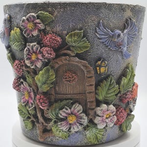 May include: A decorative planter with a textured, gray finish. The planter features raised relief details of flowers, berries, leaves, a wooden door, and a blue bird. The planter has a whimsical, fairy tale theme.