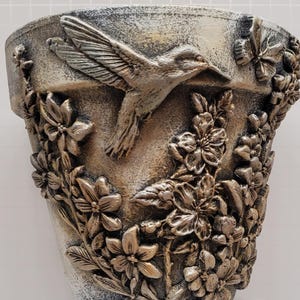 May include: A tapered plant pot with a weathered, metallic finish featuring a raised, three-dimensional relief design. The surface displays a hummingbird in flight, a butterfly, and clusters of detailed flowers and leaves wrapping around the exterior.