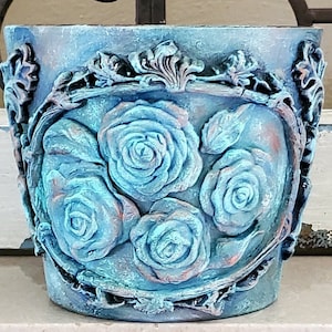 May include: A teal blue ceramic planter with a raised design of roses and vines. The planter has a distressed finish and is perfect for adding a touch of rustic charm to your home decor.