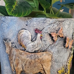 May include: A ceramic planter with a distressed white and blue finish. The planter features a rooster, a cow, and butterflies. The planter is decorated with a vintage-inspired design.