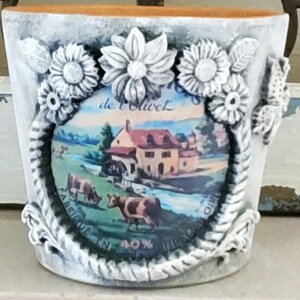 May include: A white ceramic planter with a rope-like design around the rim. The planter features a vintage-style image of a farmhouse with a windmill and cows in a field. The image is surrounded by flowers and leaves. The text "de l'Olivet" is visible on the image.