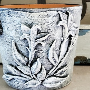 May include: White ceramic planter with a textured surface and embossed floral design. The planter has a distressed look and features a light gray and white color scheme.