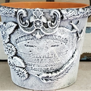 May include: White ceramic planter with a distressed finish and floral details. The planter has a label that reads "The Seville Wax & Candle Co.  Patent No. 5,784,123  Kiney Wick Works  London, E."