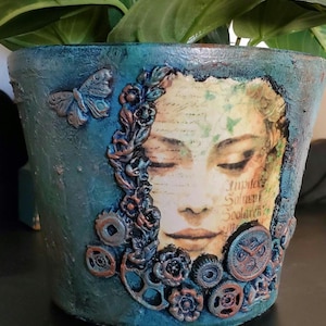 May include: A blue and green ceramic planter with a woman's face and floral design. The planter is decorated with metal gears, butterflies, and other embellishments.