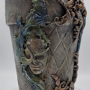 May include: A decorative gray planter with a textured finish, featuring a sculpted mask and floral accents. The planter has a diamond pattern and is adorned with green, blue, and gold details. A unique piece for home decor.