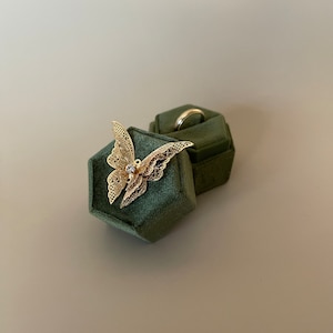May include: A gold butterfly brooch with detailed openwork, set with a small gemstone. It sits on a dark green velvet ring box, alongside a silver ring. The background is a neutral, light grey.