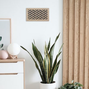 May include: A modern interior scene featuring a large potted snake plant with green and yellow striped leaves. A small wooden stool supports the plant. A wooden framed wall art with a black wave pattern hangs on the white wall.