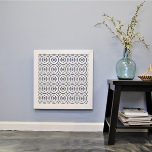May include: White decorative wall vent with a geometric pattern, mounted on a light blue wall. A small black end table with a vase of white flowers and a woven basket sits to the right of the vent.