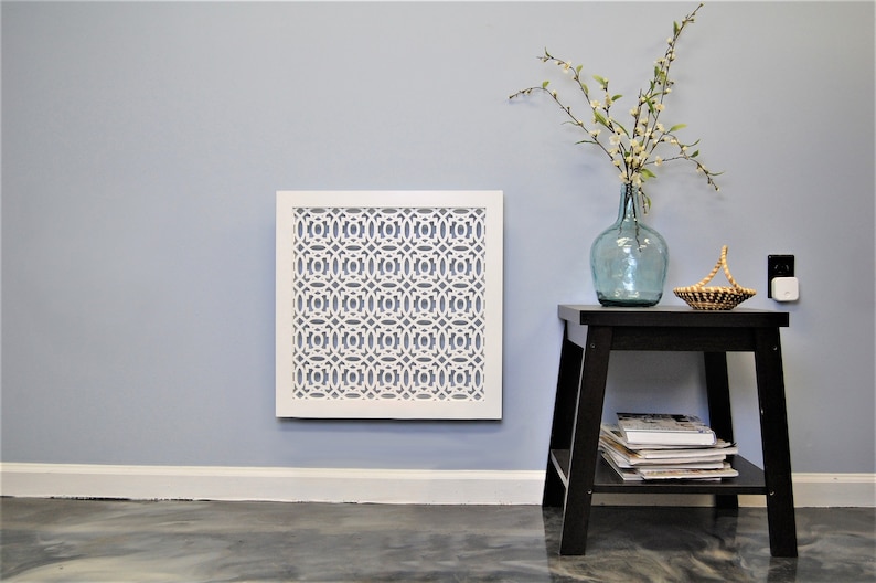 May include: White decorative wall vent with a geometric pattern, mounted on a light blue wall. A small black end table with a vase of white flowers and a woven basket sits to the right of the vent.