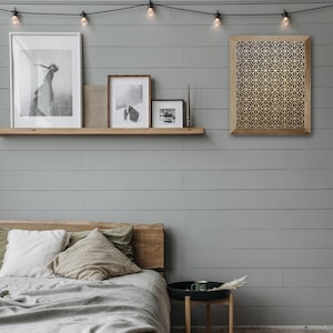 May include: A bedroom interior with a bed, a wooden shelf with framed photos, a wooden side table with a mug, and a geometric wall hanging with a wood frame.