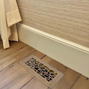 May include: A rectangular floor vent with a geometric design, set into a wooden floor. The vent cover is a light brown color with a black interior. The surrounding floorboards are also light brown, with a cream-colored wall and baseboard in the background.
