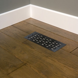 May include: A black metal floor vent with a geometric pattern of diamond shapes. The vent is rectangular and lies on a wooden floor with a dark brown finish. White baseboards and a gray wall are in the background.