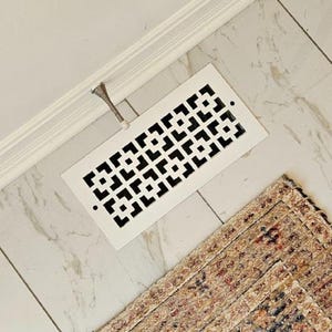 May include: A white rectangular air vent with a geometric pattern is installed on a marble tile floor. A small, decorative metal piece is attached to the vent. A patterned rug is in the lower right corner.