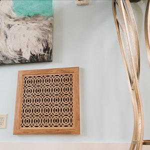 May include: A wooden wall vent cover with a decorative geometric pattern. The vent cover is light brown and has a square shape. It is mounted on a light blue wall.