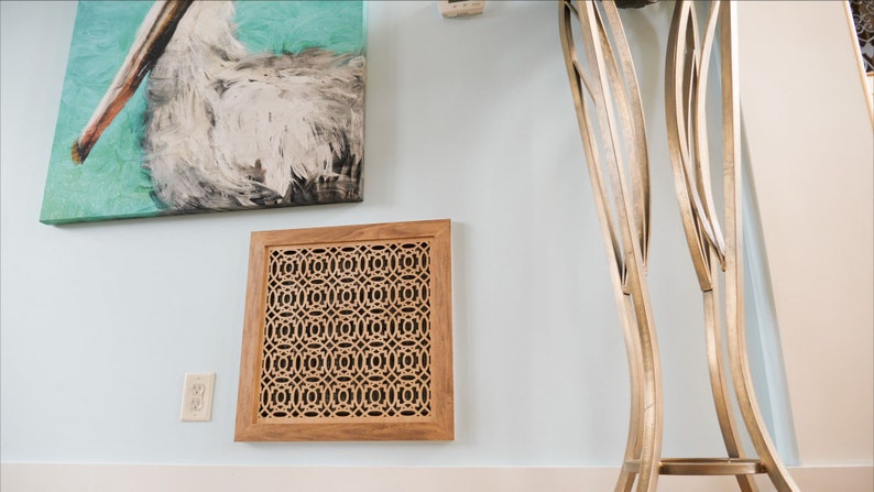 May include: A wooden wall vent cover with a decorative geometric pattern. The vent cover is light brown and has a square shape. It is mounted on a light blue wall.