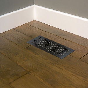 May include: A black rectangular floor vent with a decorative, openwork design. The vent is made of metal and is set into a dark wood floor. The vent is near a white baseboard and a gray wall.