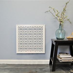 May include: White metal decorative vent cover with a geometric pattern, mounted on a light blue wall. A small black side table with a shelf holds a blue glass vase with white flowers and a woven basket.