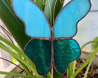 May include: A stained glass butterfly garden stake with blue and green wings. The stake is made of copper wire and is inserted into the ground.