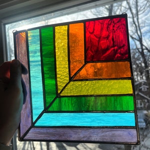 May include: A square stained glass window with a geometric design featuring a rainbow of colors, including purple, blue, green, yellow, orange, and red.