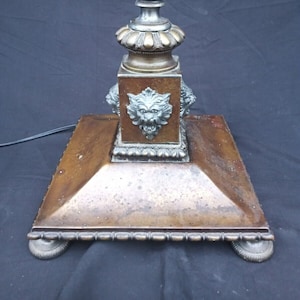 May include: A tall, antique floor lamp with a bronze base and a silver-colored top. The base features a square platform with four lion head sculptures and a decorative border. The lamp has a white shade.