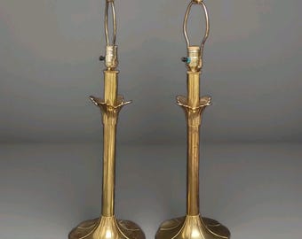May include: A pair of matching brass table lamp bases featuring a tall, slender cylindrical stem, a flared petal-shaped base, and a decorative fluted collar below the socket. Each lamp includes a metal harp and a small spherical finial.