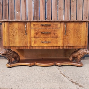 May include: A vintage wooden sideboard with a warm, natural finish. It features two cabinet doors, three drawers, and is supported by two carved lion figures. The sideboard has ornate hardware and a decorative top edge.