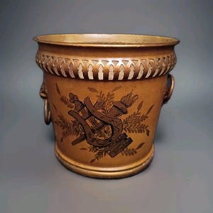 May include: A cylindrical brass bucket featuring a decorative pierced gallery rim and two side ring handles. The exterior surface displays a dark brown etched graphic of a lyre, a torch, and leafy sprigs centered on the warm metallic body.