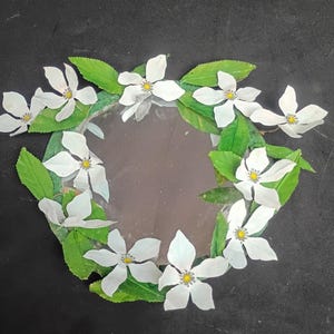 May include: A circular mirror framed by a decorative border of white five-petaled flowers with yellow centers and green serrated leaves. The flowers and leaves are arranged around the entire perimeter of the reflective glass surface.