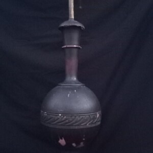 May include: An antique, dark-colored lamp with a bulbous base and a tall, slender neck. The lamp features a decorative band around the base and a brass fixture at the top. The lamp stands approximately 18 inches tall.