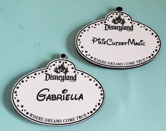 May include: Two white Disneyland name tags with black text and a black castle logo. The top tag says "Disneyland Resort Pixie Cutzof Magic Where Dreams Come True". The bottom tag says "Disneyland Resort Gabriella Where Dreams Come True".