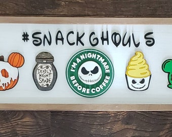 May include: A wooden sign with the text "#SNACK GHOULS" in black lettering. The sign features five Halloween-themed characters: a pumpkin with a skull face, a bottle with the text "DEADLY NIGHT SHADE", a green circle with the text "I'M A NIGHTMARE BEFORE COFFEE" and a skull face, a yellow ice cream cone with a skull face, and a green apple with a skull face.