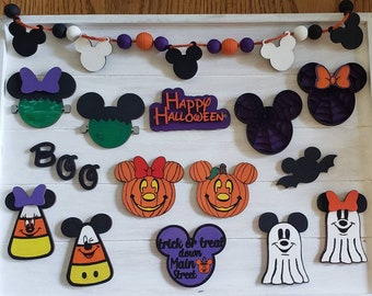May include: A collection of Halloween-themed wooden ornaments featuring Mickey Mouse and Minnie Mouse in various costumes, including a pumpkin, a ghost, a spider, and a candy corn. The ornaments are painted in bright colors and have a festive feel. The text "Happy Halloween" and "Boo" are also included.