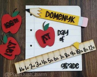May include: A wooden sign with the words "1st Day of Grade" and a ruler with numbers 1 through 12. There are also two red apples with the words "Last" and "1st" on them. The sign is decorated with a yellow pencil with the name "Domenyk" written on it.
