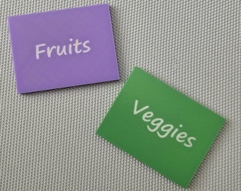 May include: Two rectangular labels, one purple and one green, with white text. The purple label reads "Fruits", and the green label reads "Veggies". The labels are on a textured, light gray surface.
