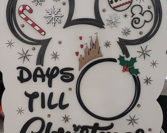 May include: White wooden sign shaped like Mickey Mouse ears with a black outline. The sign says "Days Till Christmas" with a circle in the middle for a number to be placed. The sign is decorated with snowflakes, candy canes, ornaments, and a castle.