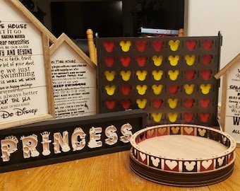 May include: A collection of wooden signs with various Disney-themed quotes, including "In this house we let it go because Hakuna Matata", "We Do Disney", "We believe in Magic", "Galaxy Far Far Away", "We Do Geek", and "Princess". There is also a wooden game board with red and yellow Mickey Mouse ears and a wooden tray with a Mickey Mouse ear design.