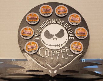 May include: A black and gray wooden coffee pod holder shaped like Jack Skellington from the movie "The Nightmare Before Christmas". The holder has the text "I'm a Nightmare Before Coffee" and features eight slots for coffee pods. The holder is decorated with a black cat and a bat.