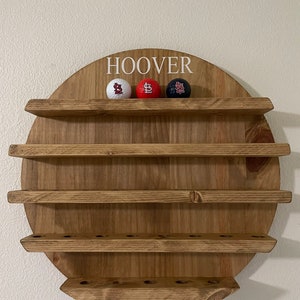 May include: A wooden wall-mounted shelf with five shelves, painted in a golden oak finish. The top shelf has the name "HOOVER" written in white lettering. Three golf balls with a baseball team logo are displayed on the top shelf.