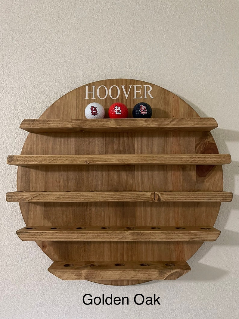 May include: A wooden wall-mounted shelf with five shelves, painted in a golden oak finish. The top shelf has the name "HOOVER" written in white lettering. Three golf balls with a baseball team logo are displayed on the top shelf.