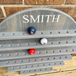 May include: A gray wooden golf ball display with the name "SMITH" written on the top. The display has multiple rows of holes to hold golf balls. There are three golf balls in the display, one red, one white, and one blue.
