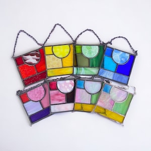 May include: Eight stained glass suncatchers with geometric designs in various colors, including red, yellow, green, blue, pink, and purple. Each suncatcher is suspended from a silver chain.
