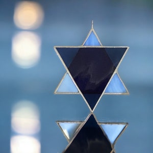 May include: A blue and silver Star of David, a six-pointed star, with a dark blue center. The star is made of stained glass and is reflecting light.