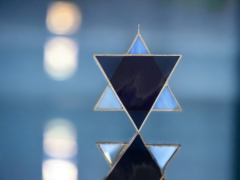 May include: A blue and silver Star of David, a six-pointed star, with a dark blue center. The star is made of stained glass and is reflecting light.