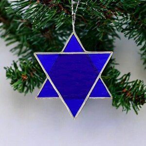 May include: A blue glass Star of David ornament with silver foil accents. The ornament is hanging from a Christmas tree branch.
