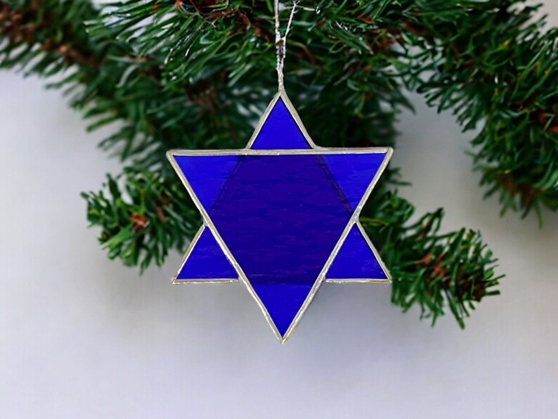 May include: A blue glass Star of David ornament with silver foil accents. The ornament is hanging from a Christmas tree branch.