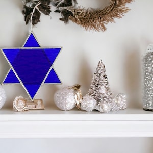 May include: A blue stained glass Star of David ornament on a white shelf with silver and white Christmas ornaments and a silver and white Christmas tree.